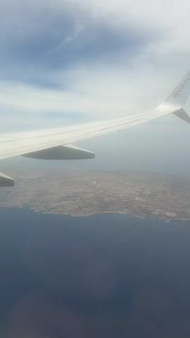 The video shows an aerial view from the window of a Ryanair airplane. The wing of the plane, with the Ryanair logo, is visible against a sky with scattered clouds. Below, the coastline of an island is visible where the land meets the deep blue sea. The island appears densely populated with buildings and dry land.