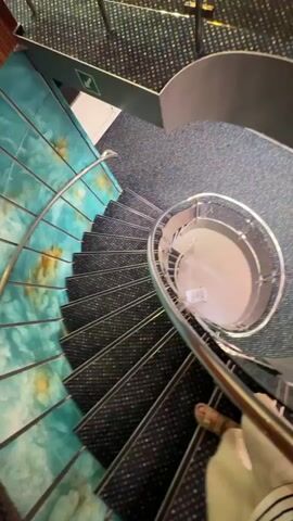 A first-person perspective video shows a person walking down a spiral staircase. The staircase has dark gray treads with a speckled pattern and a metallic railing. The wall to the left of the stairs is decorated with a turquoise and brown mosaic pattern. The person's legs and feet are visible in the lower portion of the frame, wearing light-colored pants and tan sandals. The camera angle is looking down the staircase, giving a sense of descent.