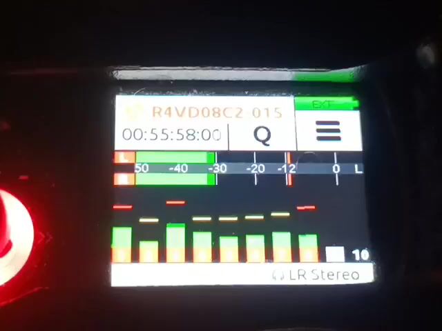 The video shows a close-up of a digital audio recorder's screen. The screen displays a recording session with a file name "R4VD08C2-015" and a timestamp that progresses from "00:55:58:12" to "00:56:01:12". There are audio level meters displayed as bar graphs, indicating that sound is being recorded. The meters show activity across different decibel levels, with some channels peaking around -12 dB and others at lower levels. The text "EXT" is visible in a green box on the top right, possibly indicating an external input is active. At the bottom of the screen, "LR Stereo" is displayed, indicating the recording format. The overall scene is dark, with only the illuminated screen and a hint of a red light on the left side of the frame being visible. The image suggests the recording is taking place in a controlled environment, likely at night or in a dimly lit area, given the prominence of the screen's backlight. There are no people, weather conditions, or explicit location cues, but the context implies an active recording process.