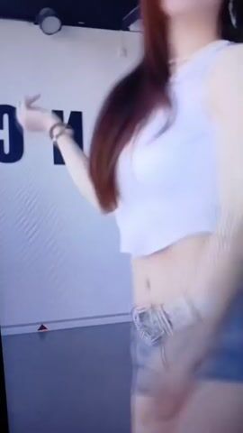 A young woman with long, dark brown hair and fair skin is shown dancing in a studio. She is wearing a white crop top, denim shorts, and a belt with a jeweled buckle. The background features a white wall with large black letters, possibly part of a studio name. The lighting is bright, and the camera is focused on the woman's midsection and upper body as she moves to the music. Her facial features are visible, and she appears to be looking towards the camera. The video is a short clip, likely from a dance performance or social media post.