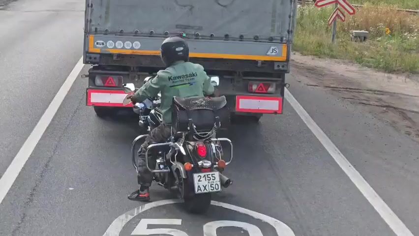 A motorcycle ridden by a person in a green jacket and helmet is positioned behind a large truck on an asphalt road. The motorcycle has saddlebags and a license plate that reads "2155 AX 50". The truck is grey and has "www.aristatrans.eu" written on its side. A railroad crossing sign with a red X and the word "СТОП" (STOP) is visible on the right side of the road, along with a smaller red and white sign. The road has white lane markings and a painted circle with the number "50" within it. The motorcycle is stopped, presumably due to the truck, and the rider briefly touches their helmet. The environment appears to be rural, with grassy areas and some trees visible in the background.