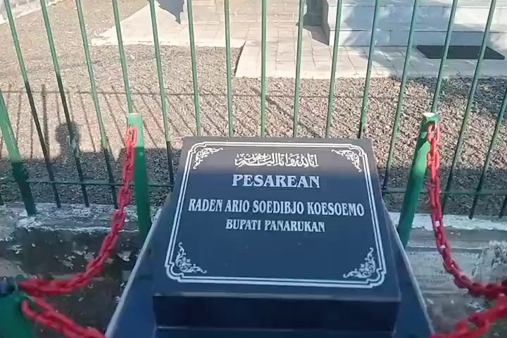 The video pans across a grave marker inscribed with Arabic script and the text "PESAREAN RADEN ARIO SOEDIBJO KOESOEMO BUPATI PANARUKAN". The grave marker is black with white lettering and decorative border. Red chains connect to green metal posts in front of the marker. Behind the grave marker is a stone structure with a roof supported by marble pillars. Steps lead up to the structure. The ground is covered in gravel. Trees and a building with a dome are visible in the background. The sky is clear and blue.