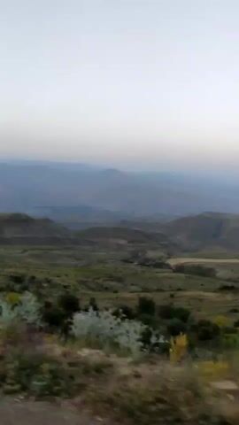The video shows a landscape from a moving vehicle. It is daytime, possibly early morning or late evening, with a clear sky and soft light. The terrain is hilly and covered with dry grass, bushes, and some trees. In the distance, there are more hills and mountains. The motion blur indicates the camera is in transit, likely on a road. There are no people or animals visible. The overall impression is of a rural, somewhat arid environment.