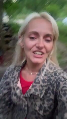 A woman with blonde hair and makeup is speaking to the camera. She is wearing a red shirt and a leopard-print jacket. The background appears to be outdoors with trees and greenery. The camera is moving, suggesting the person is walking or the camera is being held by someone walking. There is also a pink object, possibly a toy, visible in the lower left corner of the frame at 00:01 and 00:04.