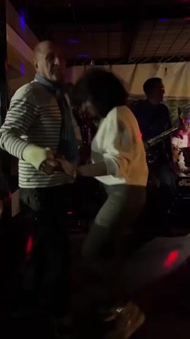 A small band plays as several people dance on a small patio. One couple consists of an older man in a striped sweater and light blue scarf, dancing with a woman in a white shirt with an open back. A guitarist and saxophone player are visible, as is a drummer. The patio is covered. A couple in leather jackets dance further back. Multi-colored lights illuminate the scene.