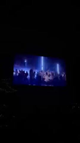 The video shows a large screen displaying a dance party. The screen is backlit and shows a crowd of people dancing and illuminated by bright blue and white lights. The screen is positioned against a dark background, with a blurred cityscape and string lights visible below.