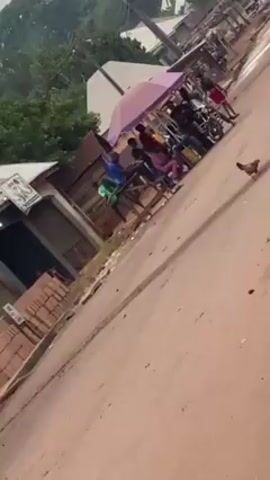 The video shows a street scene in a rural or semi-rural area. The road is unpaved and reddish in color. On the left side of the road, there are stacks of red bricks in front of a building with a sign. A group of people is seated under a pink umbrella. There are motorcycles parked nearby. In the background, there are other buildings and trees. A child in a yellow shirt and another in red run down the street, and a chicken is also present on the road. A small, white truck is parked on the side of the street at the end of the video. The sky is overcast.