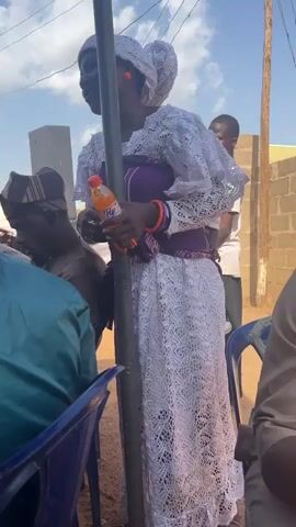 The video shows a group of people gathered outdoors under a temporary tent or structure. A woman in a white lace dress and head covering stands next to a support pole, holding an orange soda bottle. She appears to be addressing the seated group of men, gesturing with her hands. The men are dressed in traditional attire, some wearing patterned hats. There are blue plastic chairs set up for seating, and a few individuals are standing in the background. The setting looks like a community event, potentially a celebration or gathering. The sky is visible with light clouds.
