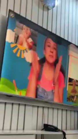 A television displays a woman in overalls clapping her hands. The screen also features two small video boxes, one with a baby clapping and another with a baby in the grass. The background of the woman's video is bright blue with a sun and clouds on one side, and a window on the other. The television sits on a white shelf, and a black landline phone is also on the shelf. The wall behind the TV is white with vertical grooves. A clock hangs on the wall above the TV.