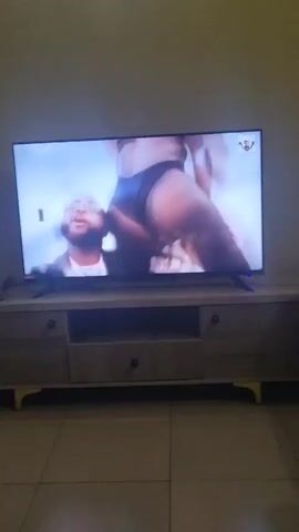 A television on a light-colored, wooden media console displays a music video. The video features two men in dark suits with white shirts and sunglasses. The man on the left has dreadlocks. The man on the right is bald and wearing glasses. The video also includes a shot of a woman in a black bikini. The room has light-colored tile floors.