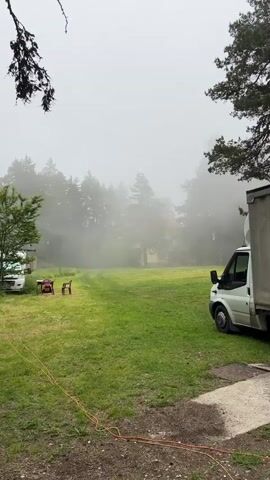 A white van is parked on the right side of the frame. A table and two chairs are situated in a grassy area on the left. A white vehicle is parked behind the table and chairs. The area is surrounded by trees and the sky is foggy.