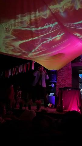 A man in a light-colored jacket and dark pants stands on a stage under a draped canopy that is illuminated with pink and yellow light. Projections of glowing green and yellow lines are displayed on the canopy. The man is holding a microphone to his mouth, as if performing. To the right of the stage, a DJ setup is visible, covered by a sparkling pink cloth. A speaker is positioned behind the DJ setup. In the background, a brick building is visible. A string of small flags hangs across the stage area. The foreground is dark, with the silhouettes of audience members visible. A laptop is on a stand near the DJ setup. The scene appears to be an outdoor nighttime event.