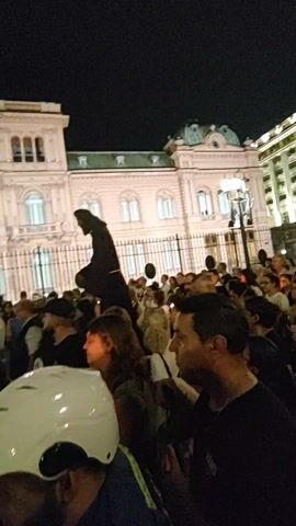 The video depicts a night-time gathering of people in front of a large, illuminated building with ornate architecture, likely a government building or palace. Many people are holding candles, indicating a vigil or procession. In the foreground, there is a person wearing a white helmet. In the center of the frame, there is a statue or figure draped in a dark robe. The sky is dark, and the overall atmosphere appears solemn and respectful.