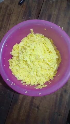 This is a close-up, overhead shot of a pile of freshly cut corn kernels in a vibrant pink bowl. The corn is a bright yellow, with individual kernels clearly visible, some appearing slightly moist. The bowl sits on a wooden surface, likely a table, with visible grain and texture. The lighting suggests an indoor setting, possibly during the day given the natural look of the corn. No people are visible in the frame. The overall impression is one of preparation for cooking or a meal. There is no visible text in the image.