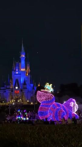 A nighttime parade float, shaped like a brightly lit, striped cat with a crown, is positioned in front of a castle. The castle is illuminated with blue and orange lights. A crowd of people are visible in the foreground, some holding glowing objects. The sky is dark.