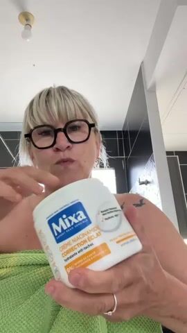 A woman with blonde hair and glasses is holding a jar of cream and applying it to her arm. She is wrapped in a green towel. The background appears to be a bathroom.