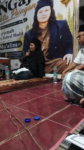 A man in a black robe and head covering speaks into a microphone while seated on a mat. He is facing a large banner featuring a picture of another man in a black suit and head covering. Two other men are seated to the right of the speaker, one facing the camera and the other looking towards the speaker. There are bottles of water on the floor in front of the speaker and a book on the floor to the right. The room has a tiled floor and the walls are not visible. The lighting appears to be artificial.