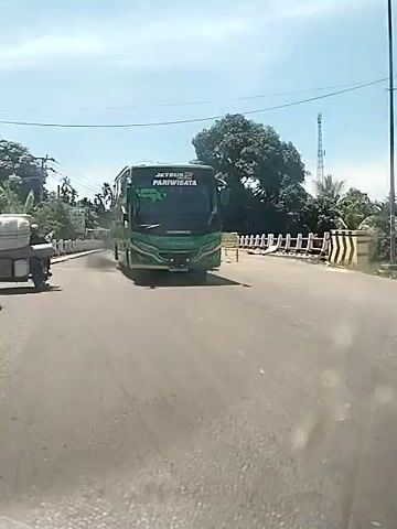 The video shows a road on a sunny day, with a bridge in the distance. A green tourist bus with the words “Jetbus Pariwisata” printed on the front passes the camera, emitting a cloud of black smoke. Following the bus is a dark-colored minivan. A motorcycle with a loaded platform is parked on the side of the road. The environment includes trees, power lines, and a cellular tower.