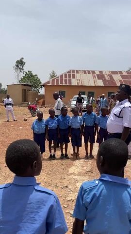 The video captures a solemn procession in Ladura Foron, Nigeria, during the daytime under clear skies.

Initially, a group of young boys, dressed in light blue uniforms with short sleeves and short trousers, are seen standing in a line on a dirt ground. They are facing an adult man in a dark uniform with a cap, who appears to be addressing them. In the background, there are simple buildings with corrugated iron roofs and a motorcycle.

The scene then transitions to a funeral procession. A wooden coffin, draped with a Nigerian flag (green, white, green), is being carried by six men in dark blue ceremonial uniforms and white gloves. They walk slowly on a dirt path. The young boys from the previous shot are now seen from behind, standing respectfully and observing the procession. Other adults are also present, with one person filming the event. A building with red-framed windows and a light-colored facade, possibly a church, is visible in the background as the procession moves past. The atmosphere is respectful and somber, indicative of a formal ceremony or funeral.