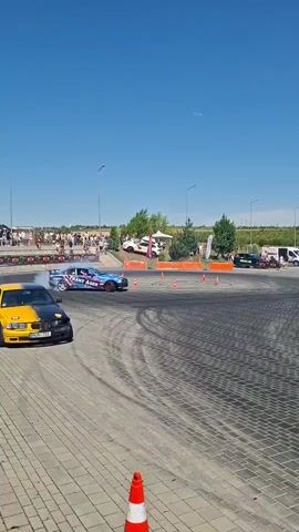The video shows a drift racing event. In the foreground, a yellow and black car is parked on a paved surface marked with tire tracks. Further back, a blue and pink race car is drifting, emitting a large cloud of smoke. Several spectators are visible in the background, along with tents and other vehicles. The sky is clear and blue.