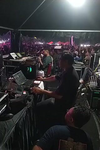 Here is a concise description of the video:

The video shows a dimly lit area behind a stage, likely during a concert or performance. There are three individuals visible: two are focused on operating sound equipment, potentially a mixing console or other audio control devices. They are crouched or leaning over the equipment, appearing to be adjusting settings or monitoring the audio output. The third individual is leaning against the equipment, seemingly tired or observing the other two. In the background, a crowd of people is visible, holding up their phones, presumably recording or taking pictures of the performance on stage. The environment is filled with various pieces of audio equipment, cables, and cases, indicating a professional sound setup.
