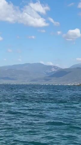 The video displays a serene seascape under a partly cloudy blue sky. The foreground is dominated by rippling, turquoise water, indicating a gentle breeze. In the mid-ground, a lush, green, tree-covered headland juts out into the water on the right side of the frame. Beyond the headland, a distant shoreline is visible with buildings suggesting a settlement. The background is characterized by a series of majestic, blue-hued mountains, their peaks softly outlined against the sky. The overall impression is one of tranquility and natural beauty. There are no people or explicit actions depicted; the video focuses solely on the landscape.