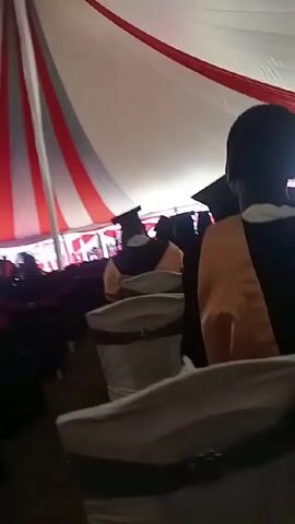 The video shows the interior of a large tent decorated with red and white fabric. A crowd of people, primarily dressed in graduation gowns and mortarboards, are seated in rows of chairs. The camera is positioned low, giving a view of the tent's ceiling and the backs of the seated individuals. A few people move around, and the overall impression is of a graduation ceremony taking place within the tent. The lighting is somewhat dim, but sufficient to discern the details of the scene.
