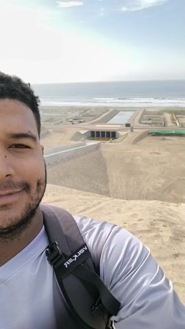 A person with dark curly hair and a short beard is facing forward. They are wearing a light grey shirt and a dark grey backpack with a white logo. They are smiling. In the background, there is a sandy landscape that slopes down to a complex of concrete structures, including what appears to be a tunnel or an overpass. Beyond the structures, there is an ocean with waves, and above that, a sky with light clouds. The wind seems strong. The shot is a close-up selfie.