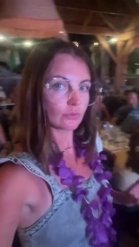 The video begins with a medium close-up of a woman with shoulder-length brown hair and glasses, wearing a grey sleeveless top and a purple lei. She is in a dimly lit indoor setting with a thatched ceiling and string lights overhead. She is looking directly at the camera and appears to be speaking or singing. In the background, there are other people seated at tables, and some are standing. The scene appears to be a restaurant or bar with a tropical theme, suggested by the lei and the decor. The woman smiles and gestures with her hands as she moves slightly, possibly dancing. The video is shaky and the focus is on the woman.