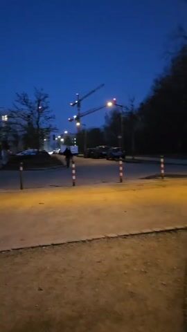 The video shows a street scene at dusk. The sky is a deep blue, and the streetlights are on, casting a yellow glow on the pavement. There are some cars parked on the side of the road and a cyclist rides by. In the background, there are two tall cranes. The camera pans slowly to the right, showing a pedestrian crossing sign and a small building. The overall scene is quiet and dimly lit.