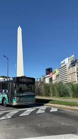 The video shows a light blue, green and black bus with “10” and other words on it driving away from the camera at a crosswalk with a large white obelisk in the background under a clear, bright blue sky. The grass in the median is green, and some buildings are visible in the background, including one with a large digital billboard on top of it.  Several people are visible near the base of the obelisk.