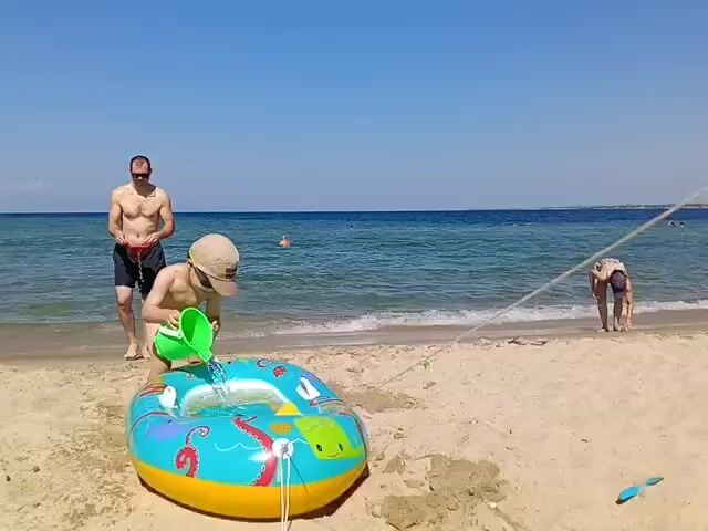 A father and son are on a sandy beach. The father, shirtless and wearing black shorts, stands a few feet behind his son. The son, wearing a hat and a light-colored t-shirt, is kneeling and pouring water from a green bucket into an inflatable pool shaped like a boat. The pool is filled with water and has an octopus and a fish design. The ocean is calm and blue, with a clear sky overhead. In the background, several other people are visible in the water and on the beach. One person is further down the beach, bending over. Another person is in the water, further out. To the left, under an umbrella, are people sitting in chairs. The scene appears to be a sunny, pleasant day at the beach.