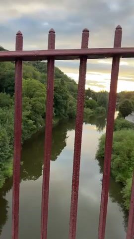 The video shows a river flowing through a valley with trees on either side. The sun is setting, casting a warm glow over the scene. In the foreground, there is a dark red metal railing, with vertical bars obscuring part of the view. On the right bank of the river, there are buildings and a road, with a few cars visible. The sky is cloudy, with patches of sunlight breaking through. The water in the river reflects the sky and the surrounding trees. The overall impression is a peaceful, natural landscape at dusk.