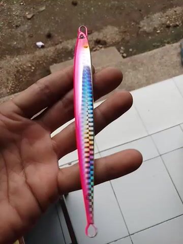 Here is a detailed description of the video:

The video shows a hand holding a fishing lure. The lure is long and thin, with a pink body and a multicolored checkered pattern on the upper side. It has metal rings at both ends. The hand holding the lure appears to be resting on a white-tiled surface. In the background, there is a concrete surface with some debris and a few stair steps. The lighting is natural, suggesting the scene is outdoors or near a window.
