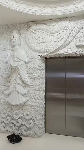 The video shows a white elevator with two doors, positioned in the center of the frame. The elevator is framed by an elaborate white bas-relief sculpture on the wall. To the left of the elevator, a sculpture of a female figure resembling a deity stands prominently. Above and to the right of the elevator, a large dragon sculpture is depicted, its head and eye visible. The wall is further adorned with intricate cloud-like patterns and other figures, creating a rich, detailed texture. The elevator control panel, with illuminated buttons, is visible on the wall to the right of the left elevator door. A small, woven stool is placed in front of the wall to the right, near the bottom of the frame. The floor appears to be light-colored. The lighting is bright, illuminating the white sculptures and the metallic elevator doors.