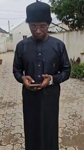 A young Black man wearing a black cap and a black traditional outfit stands outside looking at his phone. He is wearing glasses and a watch. The background shows a white building, a fence, and some plants. The ground is paved with interlocking stones. The man appears to be in a courtyard or an outdoor space.