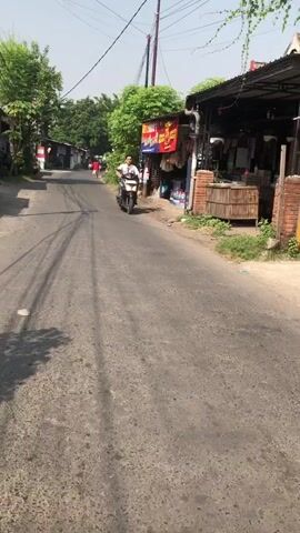 The video shows a man riding a scooter on an asphalt road. The road is lined with buildings and trees. The sky is clear and blue. The man is wearing a white shirt and black pants. He is riding the scooter at a moderate speed. The video is taken from a low angle, looking up at the man.