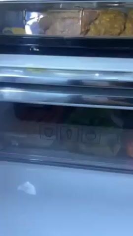 The camera shows a close-up view inside of a refrigerator. The top shelf has food containers with what appear to be fried items inside. The next shelf down is a crisper drawer with a clear front that features symbols of a bell pepper, a carrot, and a tomato. The drawer contains carrots, a bag of leafy greens, and a tomato. The next shelf down is also a clear crisper drawer with the same symbols on the front. The drawer is empty except for a small amount of condensation. The upper shelves of the refrigerator contain metallic boxes and other food containers.