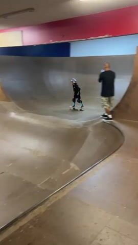 A young skateboarder in a helmet and protective gear rides up the side of a skate ramp. A man stands in the foreground, holding a phone, appearing to record the skateboarder. The skateboarder then launches into the air, performing a trick. The skate park appears to be indoors, with wooden ramps and a concrete floor. The walls are painted in red and blue. The lighting is artificial.