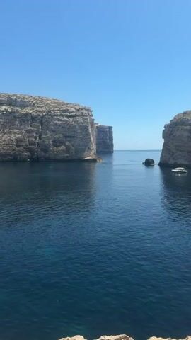 A drone captures a panoramic view of a tranquil bay on a sunny day. The deep blue water is framed by towering, rugged cliffs on either side. A sleek white yacht is anchored in the bay, with a smaller dinghy nearby. In the distance, another boat can be seen. The camera pans across the scene, revealing a person standing on the rocky shore, looking out at the ocean. The video ends with a wide shot of the vast expanse of the sea under a clear blue sky.