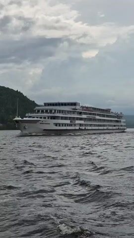 A medium shot shows a large, white multi-decked passenger ship sailing on dark, choppy water. The ship is angled slightly away from the camera, moving from left to right. The ship has many windows and balconies visible along its length. In the background, a lush green, tree-covered hill rises steeply from the water on the left side of the frame. The sky is overcast with heavy, grey clouds. The image appears to be taken from another vessel, with the edge of a window frame visible on the right side of the screen. The camera is positioned at a low angle, looking slightly upwards.