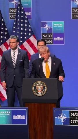 The video shows Donald Trump at a podium speaking at a NATO summit. He is wearing a dark suit and a yellow tie. Behind him stand two men, one in a suit and one in a dark blue suit. Two American flags are visible behind the speakers. The backdrop is a blue wall with "ANKARA SUMMIT NATO" branding. There are also several smaller banners on stands at the front of the stage. Trump is looking down at the podium and appears to be speaking into a microphone.