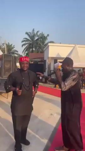 Two men in black outfits are standing on a red carpet outside a building, each holding a microphone. The man on the left is wearing a red and white knitted hat that says "CHUCK", and a black beaded necklace. He has his right hand in his pocket and a microphone in his left. The man on the right is wearing a black cap. Both men are singing or speaking into their microphones. Behind them, there are loudspeakers, a building, palm trees, and a white tent. Other people are visible in the background. The weather looks sunny.