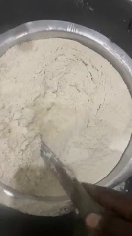A close-up overhead shot shows a metal bowl filled with a large amount of white flour. A utensil, possibly a spatula or large spoon with a wooden handle, is stirring the flour, creating swirling patterns and mounds within the bowl. The utensil is partially submerged and actively mixing the dry ingredient. The environment appears to be a kitchen or food preparation area, with a stainless steel sink visible in the background. The lighting is adequate, illuminating the flour and the mixing action.