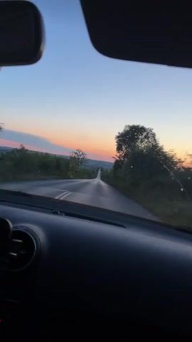 The video shows a view from inside a car driving on a road during sunset. The road stretches ahead and curves slightly. Trees and vegetation line both sides of the road. The sky is a gradient of colors, with hues of orange, pink, and blue visible near the horizon. The sun is setting, casting a warm glow on the landscape. The interior of the car, including the dashboard, is partially visible in the foreground. The camera is angled forward, capturing the road and the scenery as the car moves.