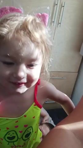 A young girl with blonde hair and a headband with sparkly pink bows smiles as she looks towards the camera. She is wearing a green swimsuit with an avocado and heart pattern. Behind her, a person's head and shoulder are visible, partially obscured by the girl. The background appears to be a room with wooden paneling and a cabinet. The lighting suggests it is daytime, possibly indoors.