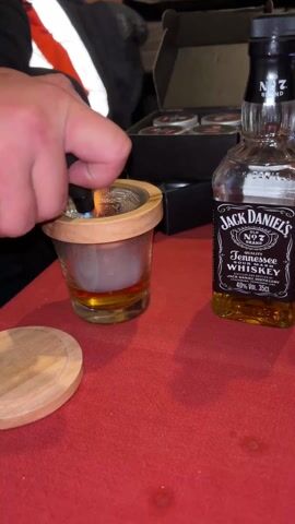 Here is a description of the video:
A person is smoking wood chips over a glass of Jack Daniels. The person uses a torch lighter to ignite the wood chips in a wooden-rimmed metal mesh strainer over the glass. The table is red, and a small, round, wooden lid is next to the glass. A box sits in the background.