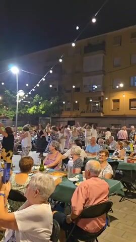 A group of people are dancing in a circle outdoors at night. They are illuminated by string lights and a street lamp. In the foreground, other people are seated at tables, some eating and drinking, while one person takes a photo with their phone. The atmosphere appears festive.