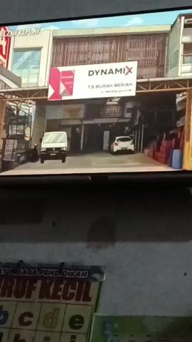 The video shows a screen displaying what appears to be a video game or animation. In the first few seconds, the scene depicts the exterior of a building with a sign that reads "DYNAMIX T.B MURAH MERIAH." There are two vehicles, a white truck and a white car, parked in front of the building. The environment looks like a commercial area with shelves and boxes visible. Then, the scene changes to an interior view of what seems to be a shop counter. A young woman with long, dark hair and wearing a white shirt stands behind the counter, looking towards the viewer with her eyes closed or downcast. The background of the shop interior shows shelves filled with various items. Below the screen, a colorful chart with letters from the alphabet and the text "HURUF KECIL" (which translates to "lowercase letters" in Indonesian) is visible.