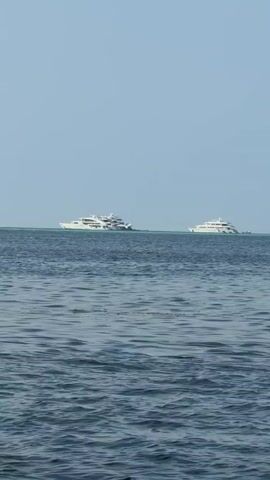 The video shows two white yachts sailing on a tranquil sea under a clear sky. The sea is calm with gentle ripples. The yachts are positioned at a distance, both parallel to each other. The background is a light blue sky.